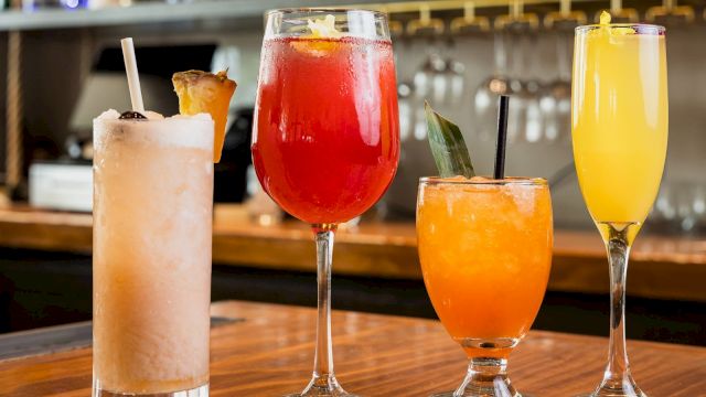 A row of colorful cocktails on a wooden bar, with bottles lined up behind the bar.
