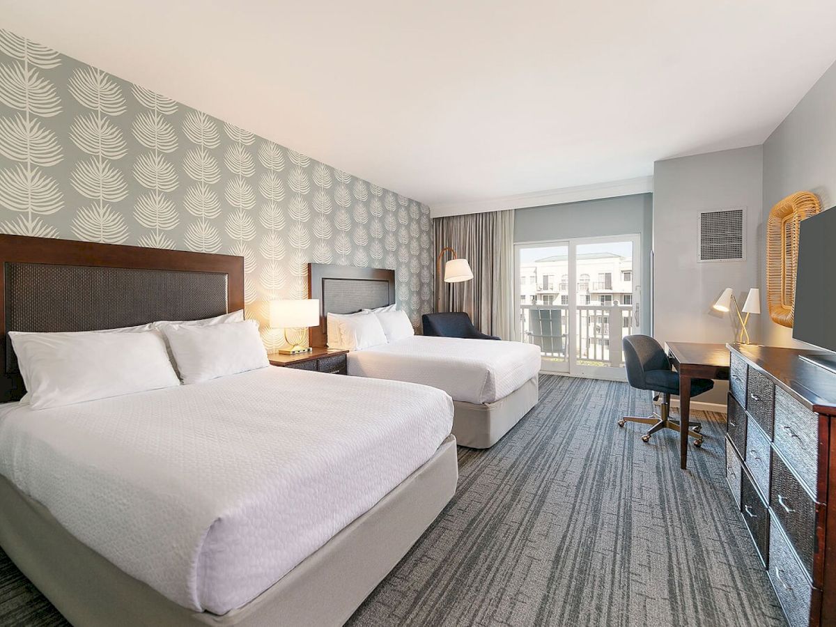 A hotel room with two queen beds, patterned wallpaper, a desk, a balcony view, and a large TV across from a dresser.
