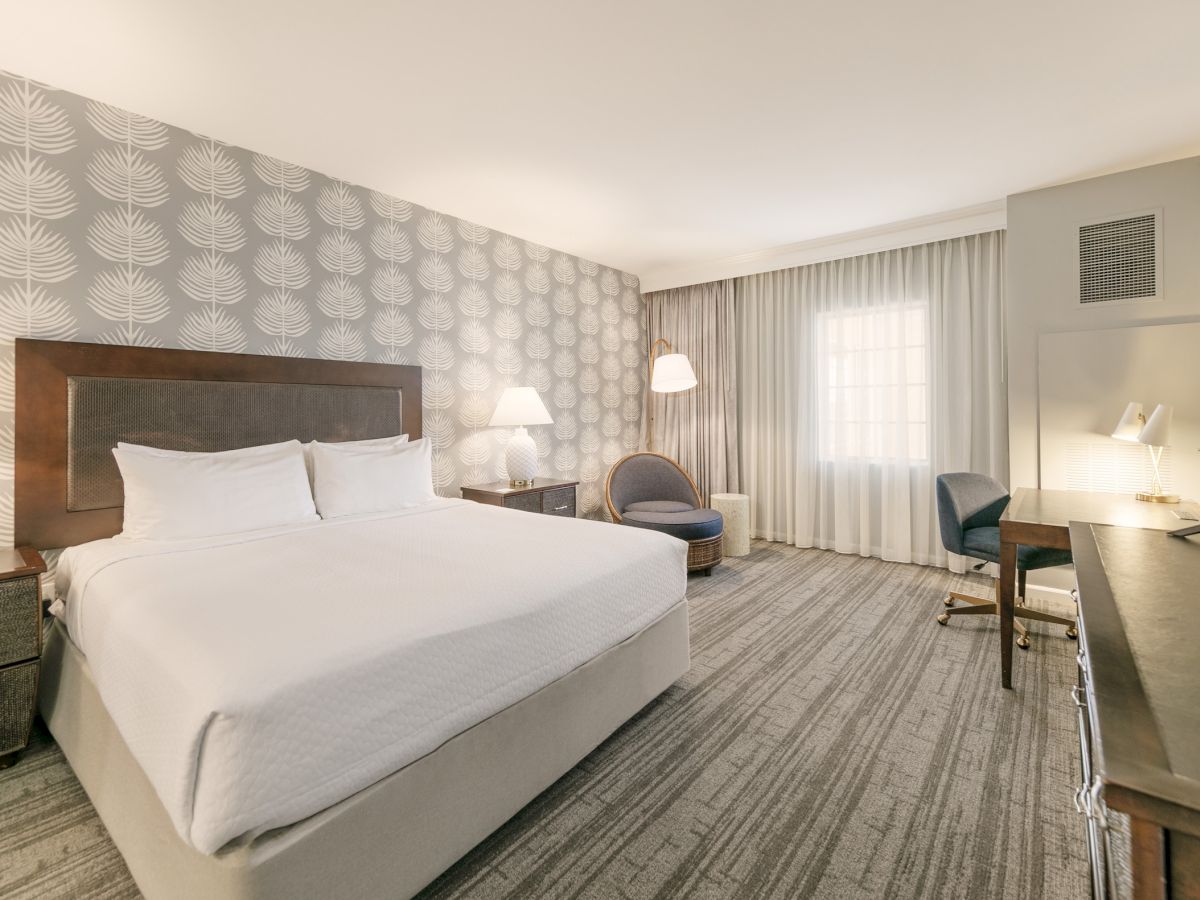 A modern hotel room with a large white bed, patterned accent wall, bedside lamps, a seating area, desk, and soft neutral decor.