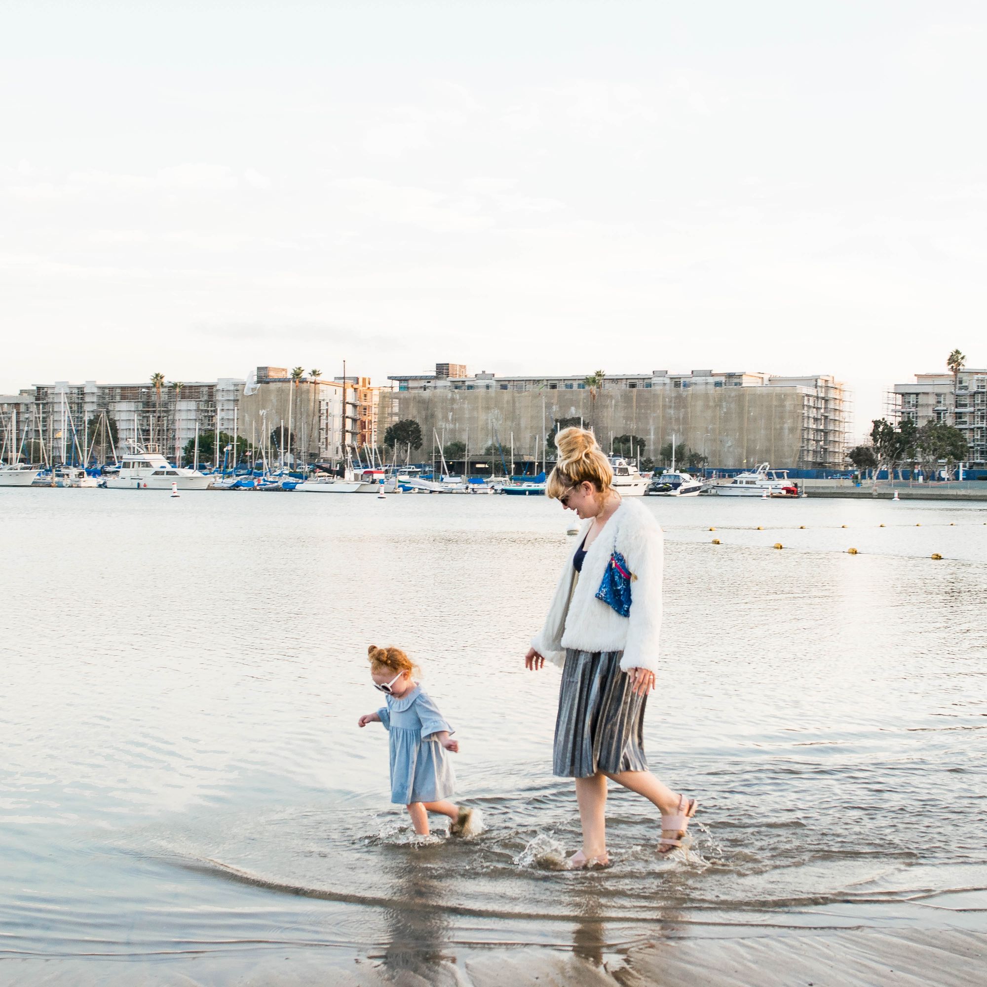 Two people walk along the shallow shoreline at a harbor, a child in blue and an adult guiding them, with boats and buildings in the background.