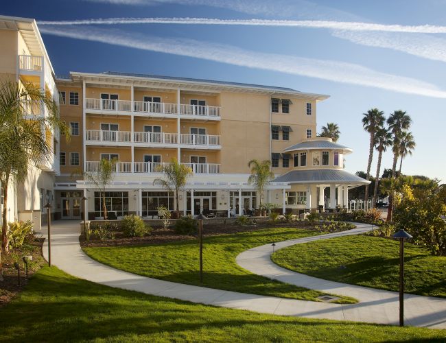 A sunny yellow hotel or resort with multiple balconies, palm trees, a curved walkway, and manicured lawns under a clear blue sky.