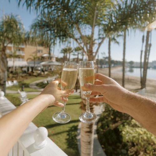 Two people are toasting with champagne glasses on a sunny balcony overlooking a tropical garden and the beach.