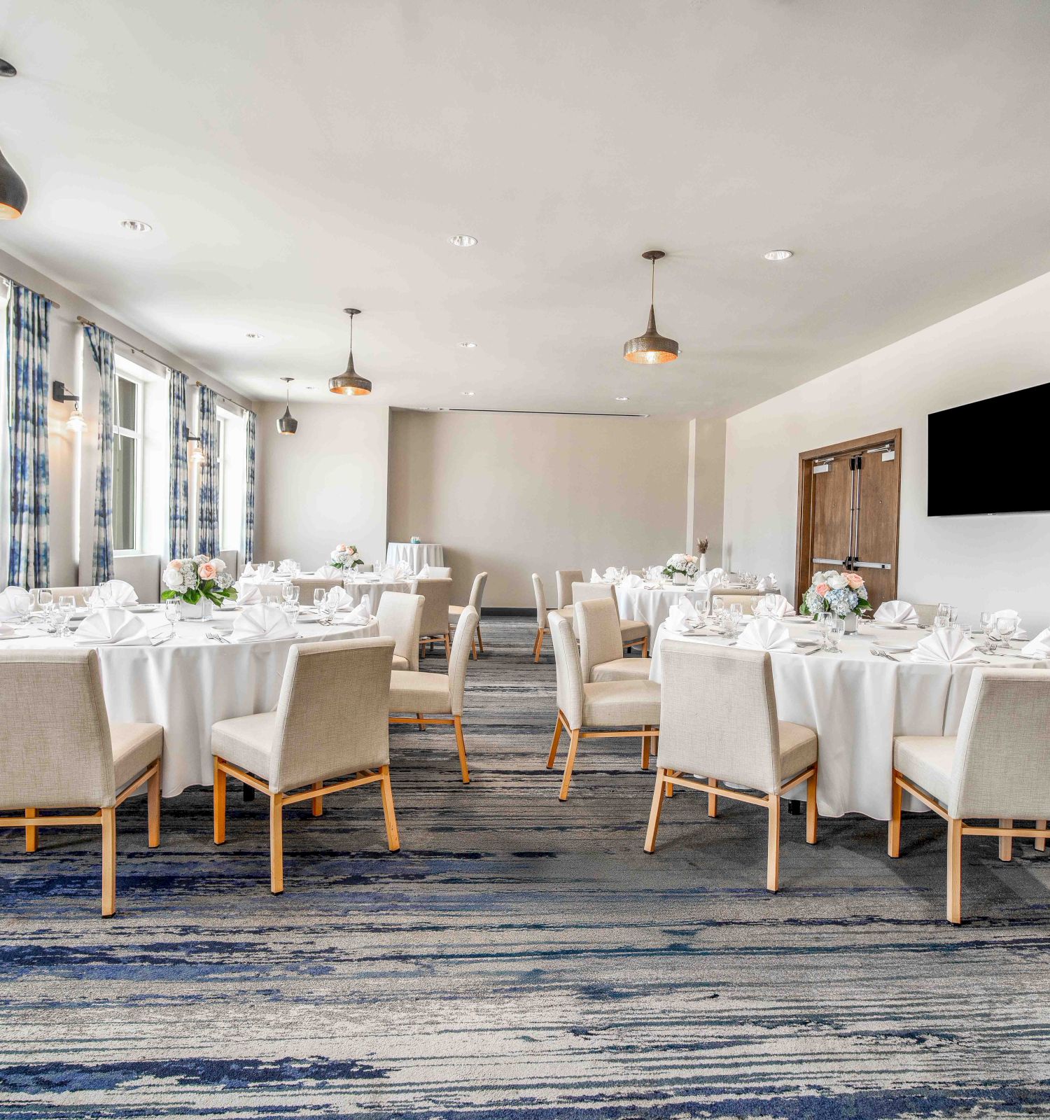 A bright, airy dining room set for a formal event with multiple round tables, white chairs, and table settings, plus a wall-mounted TV and blue-toned carpet.