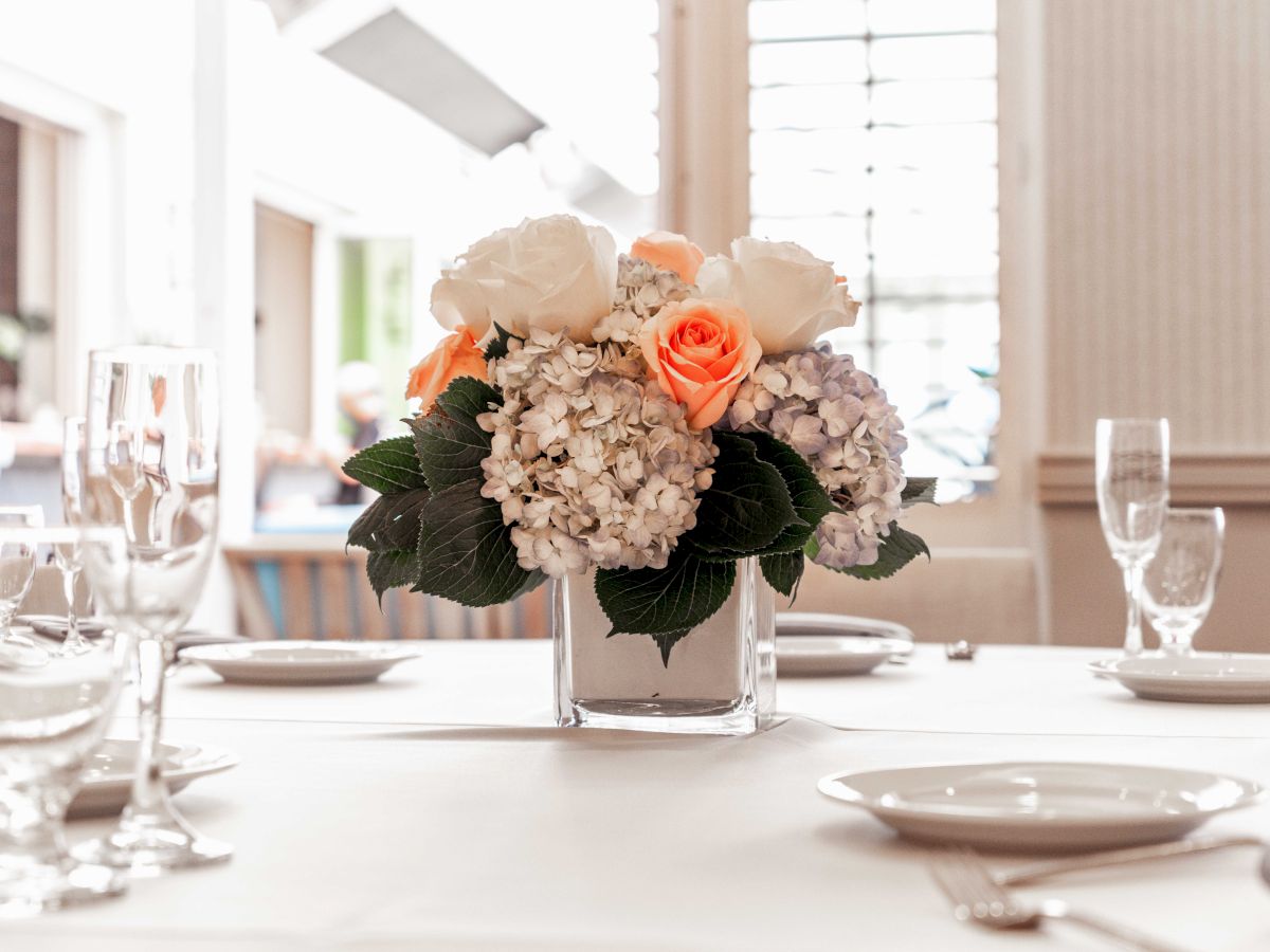 A elegant dining table setup with a white tablecloth, glassware, and a centerpiece of peach roses and hydrangeas in a white vase.