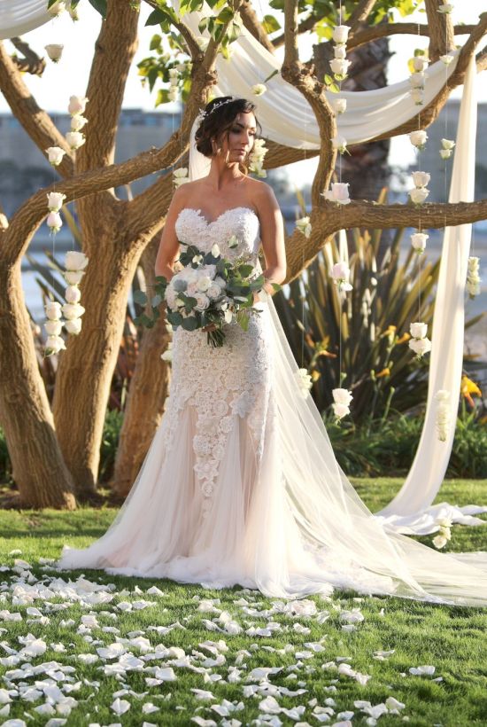 A bride stands in a flowing wedding gown under a tree, bouquet in hand, petals scattered on the grass by an archway with draped fabric.