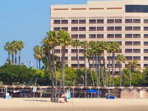 A sandy beach in front of a tall modern building, lined with palm trees, beach tents, and people enjoying the shore.