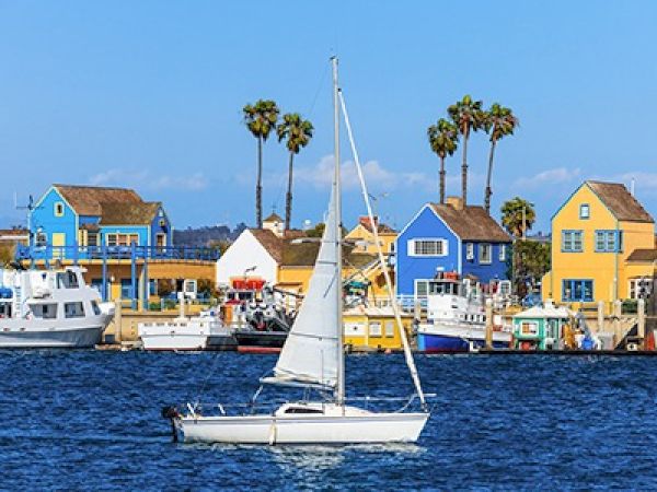 A sailboat on calm water with colorful coastal houses and palm trees in the background, a sunny seaside scene.