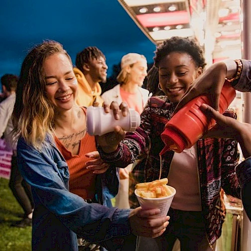 A group of friends sharing funnel cake and drinks at a fair, smiling and having fun together, with one pouring into cups.