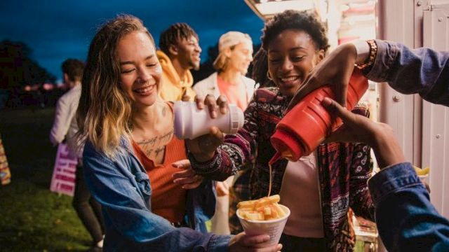 A group of friends sharing funnel cake and drinks at a fair, smiling and having fun together, with one pouring into cups.