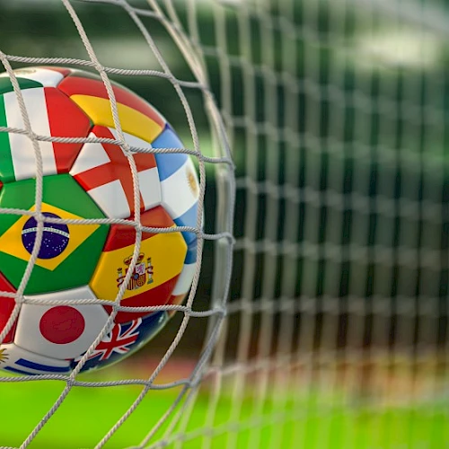 A close-up soccer ball, covered in international flags, stuck in a goal net on a grassy field.