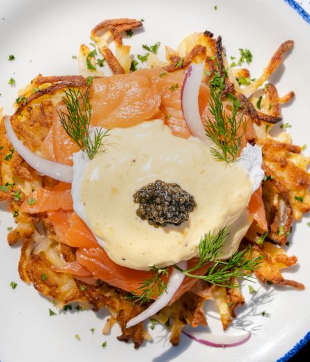 A plate of crispy potatoes topped with smoked salmon, onion slices, dill, a creamy sauce, and a dollop of caviar on top.